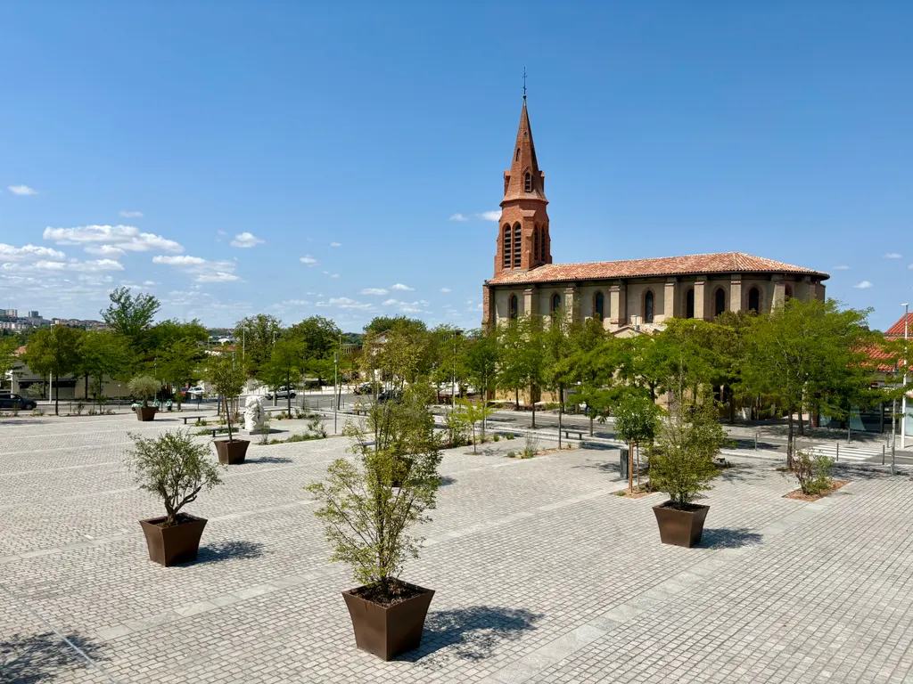 L'église de Balma et sa place pavée plantée d'arbres, sous un ciel d'été